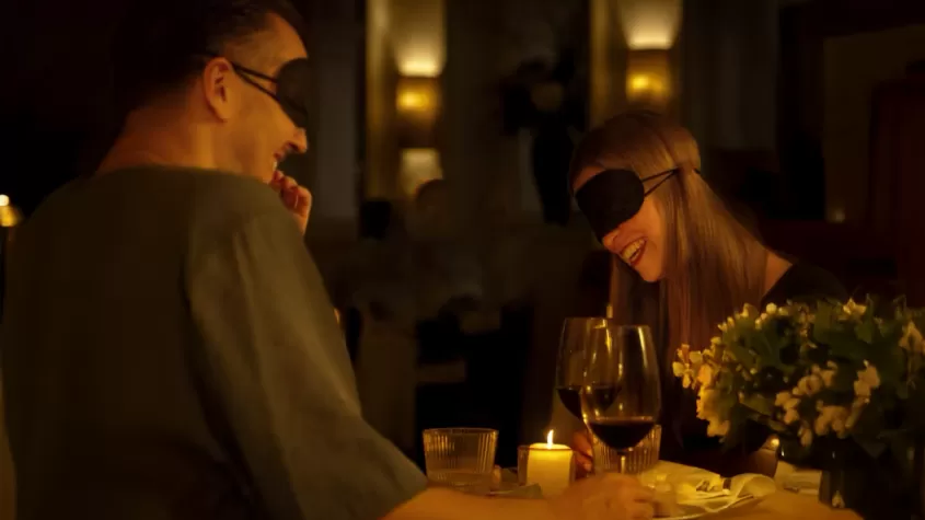 A man and a woman, both wearing black blindfolds, share a laugh while sitting at a candlelit dinner table in a dimly lit restaurant. The table is set with wine glasses, a small lit candle, and a bouquet of white flowers. The atmosphere appears intimate and elegant.