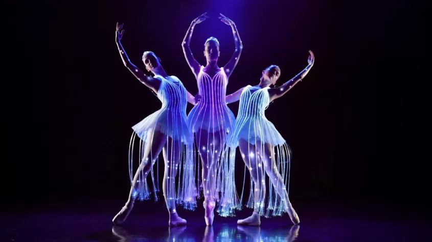 Three ballet dancers perform on a dark stage, wearing illuminated dresses with glowing fiber optic strands. The central dancer stands en pointe with arms raised, while the two dancers on either side extend their arms gracefully. The soft blue and purple lighting enhances the ethereal effect of their glowing costumes.