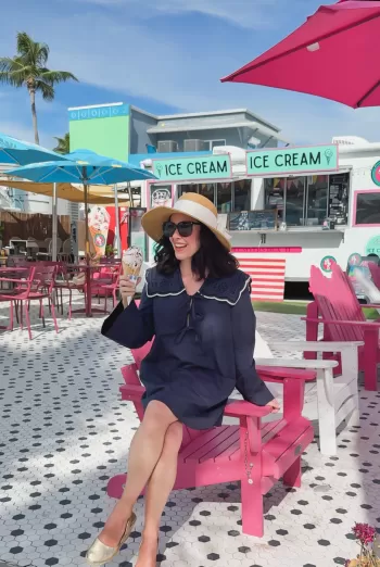 Woman in a wide-brimmed straw hat and navy dress with a sailor collar seated on a bright pink Adirondack chair eating ice cream in front of a pastel ice-cream truck.